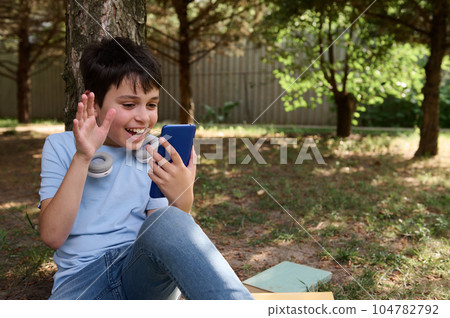 Cheerful teen boy waving hello with hand, having online conference by video link on his smartphone, sitting under a tree 104782792