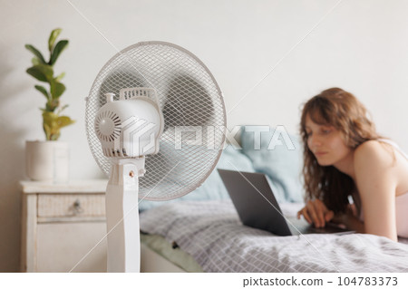 European woman cools down home with help of air conditioner fan, works with laptop in home office European woman cools down home with help of air conditioner fan, works with laptop in home office 104783373