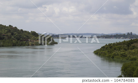 A large river with green trees on the left and right ended into a lake with a mountain background in the horizon with bright cloudy sky. 104784085