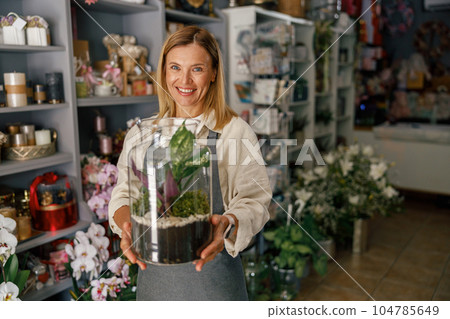 Smiling woman florist business owner standing with houseplant on background of floral store Smiling woman florist business owner standing with houseplant on background of floral store 104785649