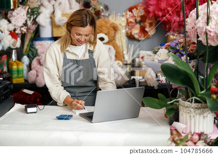 Small flower shop owner florist wearing apron working on laptop and taking online orders in store 104785666
