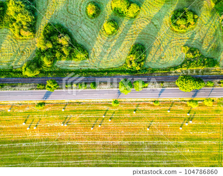 Asphalt road and concurrent railway in lush green morning lagricultural landscape. Aerial view from drone. Asphalt road and concurrent railway in lush green morning lagricultural landscape. Aerial view from drone. 104786860