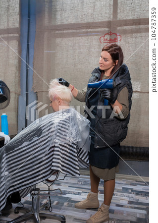 An adult female client sits in a chair in a salon in the process of hairdressing services, cutting and dyeing hair, working as a barber An adult female client sits in a chair in a salon in the process of hairdressing services, cutting and dyeing hair, working as a barber 104787359