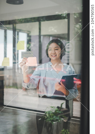 Smiling businesswoman sticking adhesive notes on glass window in office. 104790565