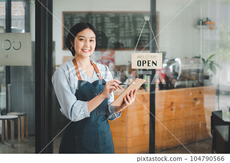 Beautiful young barista woman in apron holding order paper and standing in front of the door of cafe with open sign board. Business owner startup concept. Beautiful young barista woman in apron holding order paper and standing in front of the door of cafe with open sign board. Business owner startup concept. 104790566