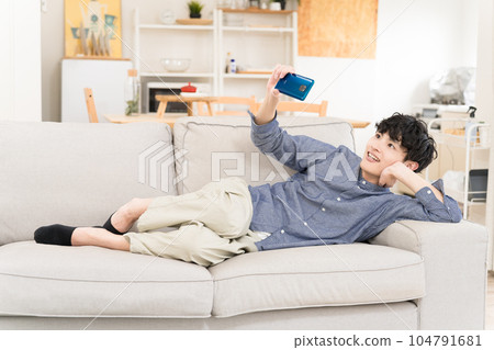 A young man lying on the sofa watching a video on his smartphone with a smile A young man lying on the sofa watching a video on his smartphone with a smile 104791681