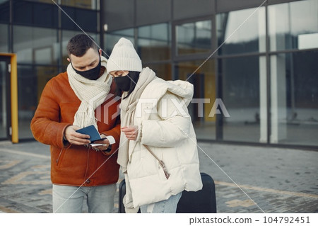 Young family going on vacation. Man and woman standing with luggage and wearing black face mask. Couple wearing warm clothes. 104792451