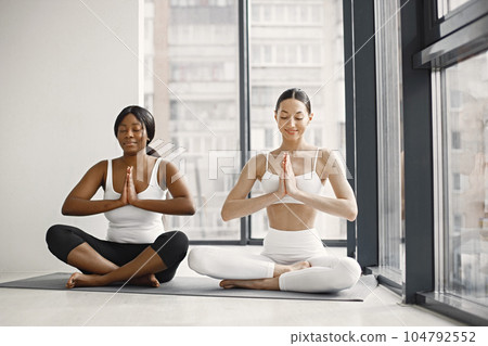 Two women stretching on yoga mat in bright studio with big windows. Caucasian woman wearing white leggins and top and black woman white t-shirt and black leggins. Sport, fitness, diet, nutrition and 104792552