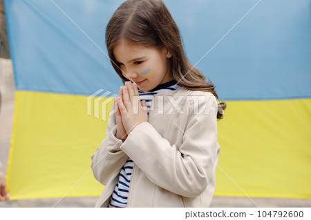 Little girl with ukrainian flag on a cheek standing in front of big flag of Ukraine. Young pretty girl praying. Girl supporting Ukraine. Little girl with ukrainian flag on a cheek standing in front of big flag of Ukraine. Young pretty girl praying. Girl supporting Ukraine. 104792600