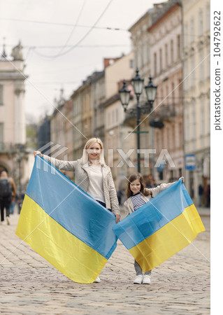 Young mother and her daughter standing on the street of old town. Girl and woman holding big flags of Ukraine in blue and yellow colours. Woman and girl supporting Ukraine against war. Young mother and her daughter standing on the street of old town. Girl and woman holding big flags of Ukraine in blue and yellow colours. Woman and girl supporting Ukraine against war. 104792622