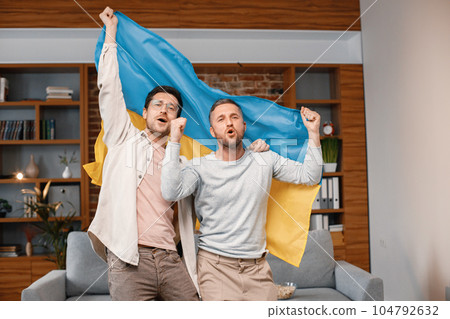 Male friends gathered at home to watch a football match. Two men standing and holding a big flag of Ukraine in hands. Men actively support their favorite team and feeling satisfied. 104792632