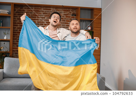 Male friends gathered at home to watch a football match. Two men standing and holding a big flag of Ukraine in hands. Men actively support their favorite team and feeling satisfied. 104792633