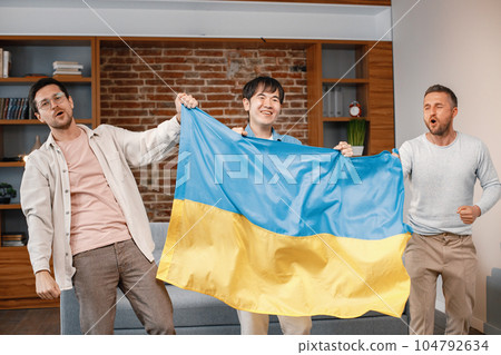 Male friends gathered at home to watch a football match. Men standing and holding a big flag of Ukraine in hands. Men actively support their favorite team and feeling satisfied. 104792634