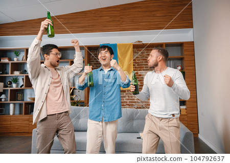 Male friends gathered at home to watch a football match. Men standing in front of big flag of Ukraine on a wall. Men actively support their favorite team and feeling satisfied. 104792637