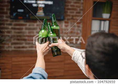 Focus on a bottles of beer. Back view of male friends gathered at home to watch a football match. Men actively support their favorite team and comment on the match 104792650