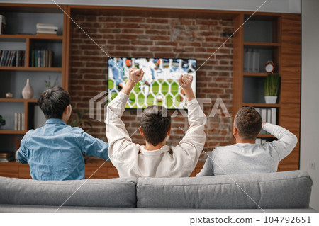 Back view of male friends gathered at home to watch a football match. Men sitting on the couch in front of the big screen TV. Men actively support their favorite team and comment on the match Back view of male friends gathered at home to watch a football match. Men sitting on the couch in front of the big screen TV. Men actively support their favorite team and comment on the match 104792651