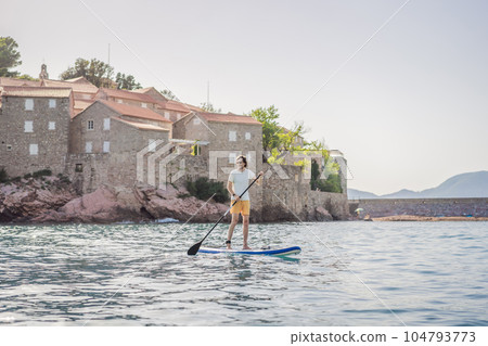 Young men Having Fun Stand Up Paddling in blue water sea near st stefan island in Montenegro. SUP Young men Having Fun Stand Up Paddling in blue water sea near st stefan island in Montenegro. SUP 104793773