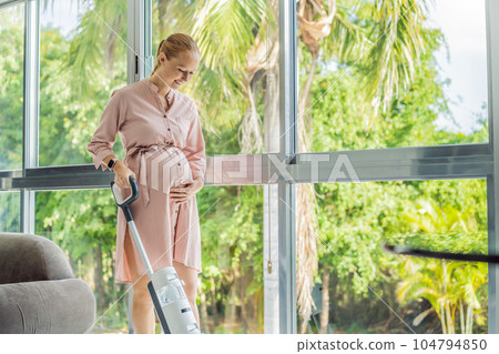 Young pregnant woman enjoys cleaning her house. Easy cleaning with a wireless vacuum cleaner. Pregnant woman cleaning floor with handheld vacuum cleaner. Modern easy cleaning Young pregnant woman enjoys cleaning her house. Easy cleaning with a wireless vacuum cleaner. Pregnant woman cleaning floor with handheld vacuum cleaner. Modern easy cleaning 104794850