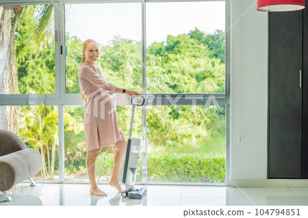 Young pregnant woman enjoys cleaning her house. Easy cleaning with a wireless vacuum cleaner. Pregnant woman cleaning floor with handheld vacuum cleaner. Modern easy cleaning 104794851