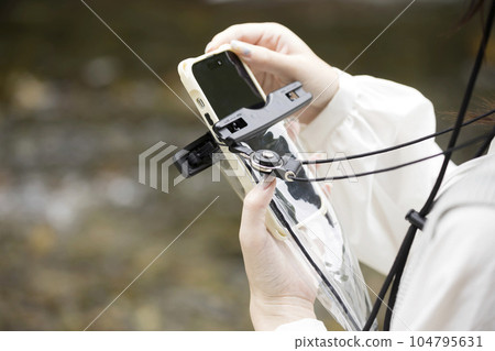 A young woman putting her smartphone into a waterproof smartphone case on the riverbank 104795631