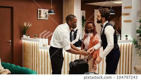 Happy african american tourists checking out from hotel after handing over room access card key to asian receptionist. Joyful guests tipping helpful bellboy carrying their luggage 104797243