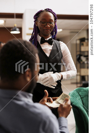 Tips and gratuities. Happy African American waitress wearing uniform receiving tip from satisfied generous customer at hotel bar, feeling excited. Traveler paying with cash for resort services Tips and gratuities. Happy African American waitress wearing uniform receiving tip from satisfied generous customer at hotel bar, feeling excited. Traveler paying with cash for resort services 104797298