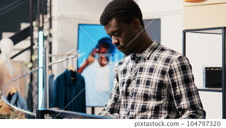 African american man shopper looking at new fashion collection, analyzing merchandise fabric in modern boutique. Shopaholic customer shopping for fashionable clothes in clothing store African american man shopper looking at new fashion collection, analyzing merchandise fabric in modern boutique. Shopaholic customer shopping for fashionable clothes in clothing store 104797320