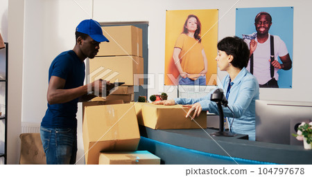 African american deliveryman asking worker to sign distribution report on tablet computer, discussing customer shipping detalis in shopping mall. Employee preparing packages in modern boutique African american deliveryman asking worker to sign distribution report on tablet computer, discussing customer shipping detalis in shopping mall. Employee preparing packages in modern boutique 104797678