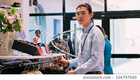 Asian worker arranging hangers with trendy clothes, working at clothing store visual. Cheerful manager preparing modern boutique store for opening, checking racks full with new fashion collection 104797680