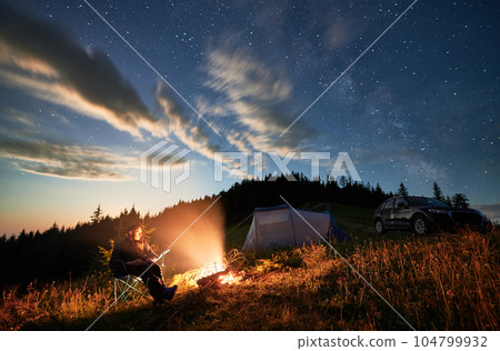 Camping in mountains under evening cloudy sky. Young man having a rest next to campfire, near tourist tent and off-road car on hill. Travelling man sitting and stargazing at night. Camping in mountains under evening cloudy sky. Young man having a rest next to campfire, near tourist tent and off-road car on hill. Travelling man sitting and stargazing at night. 104799932