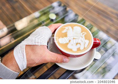Man wrists tied with an elastic bandage. Cup of hot cappuccino. The table in the coworking space of the coffee shop. A cup of coffee on the veranda on a sunny day. Coffee break at the cafe. Close-up Man wrists tied with an elastic bandage. Cup of hot cappuccino. The table in the coworking space of the coffee shop. A cup of coffee on the veranda on a sunny day. Coffee break at the cafe. Close-up 104801046