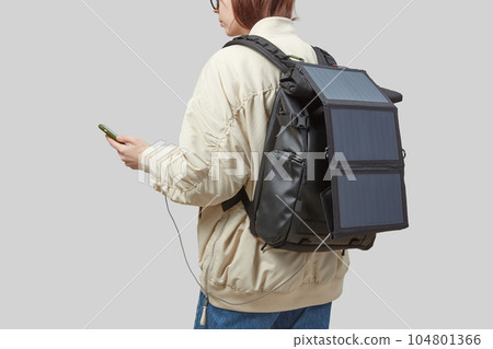 Young woman carrying backpack with flexible solar panel attached to smartphone in her hands 104801366