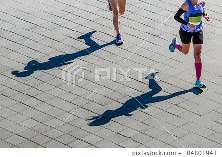overhead view two female athletes running marathon race, shadows of runners on paving slabs 104801941