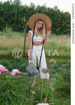 A blonde woman is watering flowers in the garden in the courtyard of a village house. A blonde woman is watering flowers in the garden in the courtyard of a village house. 104802013