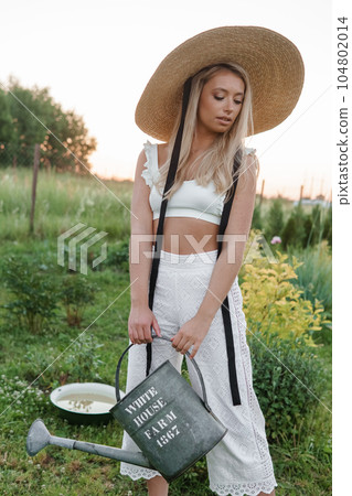 A blonde woman is watering flowers in the garden in the courtyard of a village house. 104802014