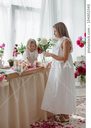 A little blonde girl with her mom on a kitchen countertop decorated with peonies. The concept of the relationship between mother and daughter. Spring atmosphere. 104802048