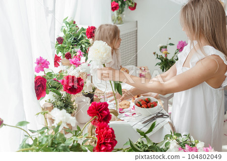 A little blonde girl with her mom on a kitchen countertop decorated with peonies. The concept of the relationship between mother and daughter. Spring atmosphere. A little blonde girl with her mom on a kitchen countertop decorated with peonies. The concept of the relationship between mother and daughter. Spring atmosphere. 104802049