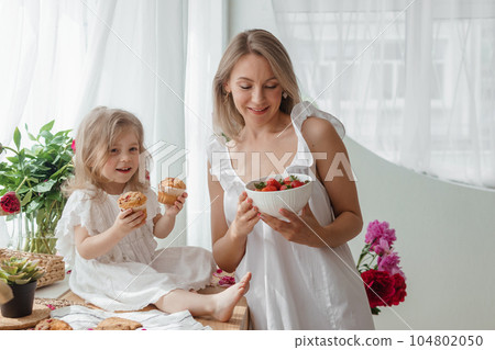 A little blonde girl with her mom on a kitchen countertop decorated with peonies. The concept of the relationship between mother and daughter. Spring atmosphere. A little blonde girl with her mom on a kitchen countertop decorated with peonies. The concept of the relationship between mother and daughter. Spring atmosphere. 104802050