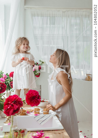 A little blonde girl with her mom on a kitchen countertop decorated with peonies. The concept of the relationship between mother and daughter. Spring atmosphere. A little blonde girl with her mom on a kitchen countertop decorated with peonies. The concept of the relationship between mother and daughter. Spring atmosphere. 104802052