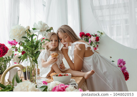 A little blonde girl with her mom on a kitchen countertop decorated with peonies. The concept of the relationship between mother and daughter. Spring atmosphere. 104802053