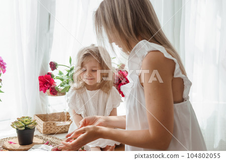 A little blonde girl with her mom on a kitchen countertop decorated with peonies. The concept of the relationship between mother and daughter. Spring atmosphere. A little blonde girl with her mom on a kitchen countertop decorated with peonies. The concept of the relationship between mother and daughter. Spring atmosphere. 104802055