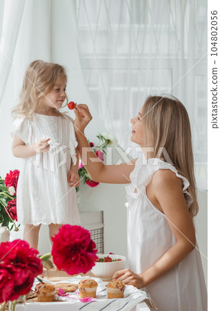 A little blonde girl with her mom on a kitchen countertop decorated with peonies. The concept of the relationship between mother and daughter. Spring atmosphere. A little blonde girl with her mom on a kitchen countertop decorated with peonies. The concept of the relationship between mother and daughter. Spring atmosphere. 104802056