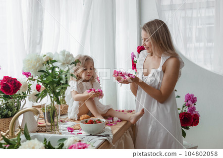A little blonde girl with her mom on a kitchen countertop decorated with peonies. The concept of the relationship between mother and daughter. Spring atmosphere. A little blonde girl with her mom on a kitchen countertop decorated with peonies. The concept of the relationship between mother and daughter. Spring atmosphere. 104802058
