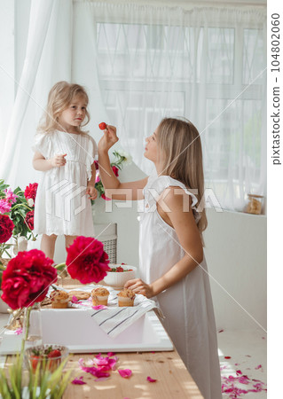 A little blonde girl with her mom on a kitchen countertop decorated with peonies. The concept of the relationship between mother and daughter. Spring atmosphere. A little blonde girl with her mom on a kitchen countertop decorated with peonies. The concept of the relationship between mother and daughter. Spring atmosphere. 104802060