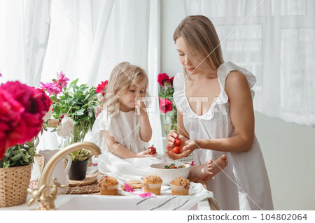 A little blonde girl with her mom on a kitchen countertop decorated with peonies. The concept of the relationship between mother and daughter. Spring atmosphere. A little blonde girl with her mom on a kitchen countertop decorated with peonies. The concept of the relationship between mother and daughter. Spring atmosphere. 104802064