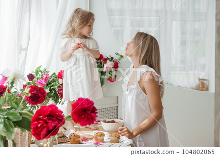 A little blonde girl with her mom on a kitchen countertop decorated with peonies. The concept of the relationship between mother and daughter. Spring atmosphere. A little blonde girl with her mom on a kitchen countertop decorated with peonies. The concept of the relationship between mother and daughter. Spring atmosphere. 104802065