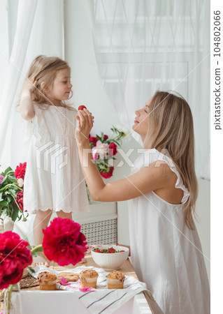 A little blonde girl with her mom on a kitchen countertop decorated with peonies. The concept of the relationship between mother and daughter. Spring atmosphere. A little blonde girl with her mom on a kitchen countertop decorated with peonies. The concept of the relationship between mother and daughter. Spring atmosphere. 104802066