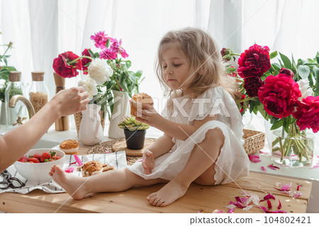 A little blonde girl on a kitchen countertop decorated with peonies. Spring atmosphere. 104802421