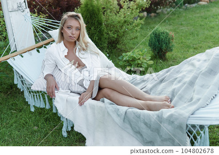 A beautiful blonde woman is lying on a hammock in her summer cottage. The concept of life in the country and summer holidays. A beautiful blonde woman is lying on a hammock in her summer cottage. The concept of life in the country and summer holidays. 104802678