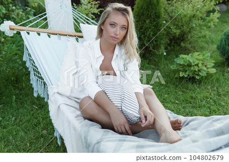 A beautiful blonde woman is lying on a hammock in her summer cottage. The concept of life in the country and summer holidays. A beautiful blonde woman is lying on a hammock in her summer cottage. The concept of life in the country and summer holidays. 104802679